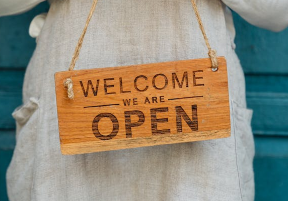 a wood sign reading "welcome we are open" held in front of an aproned body