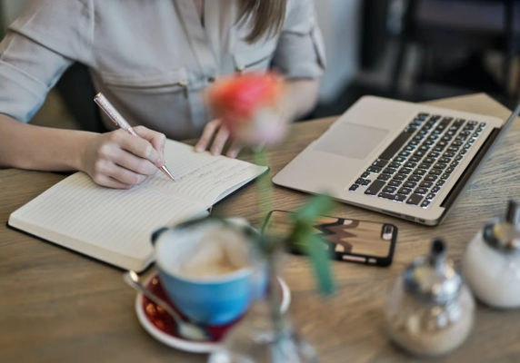 Cropped image of a white woman writing in a notebook on a table with a laptop, vase, and mug; small business writing and editing services